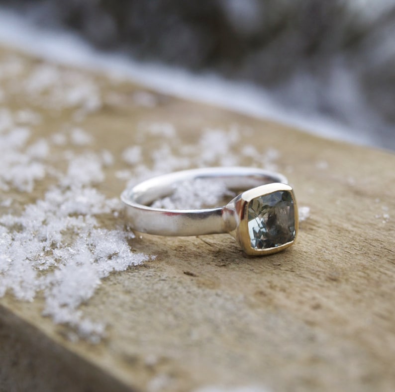 Puede incluir: Un anillo de plata con una piedra preciosa de aguamarina de forma cuadrada engastada en un engaste de oro. El anillo est&aacute; sobre una superficie de madera con nieve.