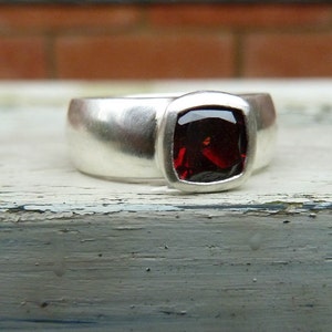 May include: A silver ring featuring a red gemstone set in a square bezel. The ring is wide and has a smooth, polished finish.