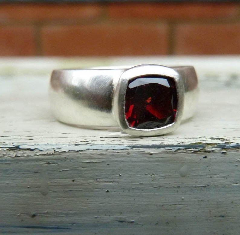 May include: A silver ring featuring a red gemstone set in a square bezel. The ring is wide and has a smooth, polished finish.