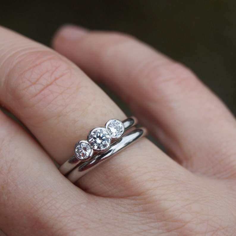 Pode incluir: Um anel de prata com tr&ecirc;s diamantes redondos engastados em bezel. O anel &eacute; usado no dedo de uma pessoa.