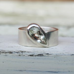 May include: A silver ring with a pear-shaped clear gemstone set in the band.
