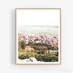 May include: A watercolour print depicting a cityscape with a body of water in the background. The artwork shows a city with buildings and trees, framed in a light wood frame. The colour palette includes shades of green, pink, and white.