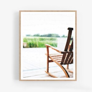 May include: A watercolour painting of a wooden rocking chair on a porch. The chair is made of light-coloured wood with a dark brown backrest. The background features a blurred green field and a bright sky, creating a serene outdoor scene.