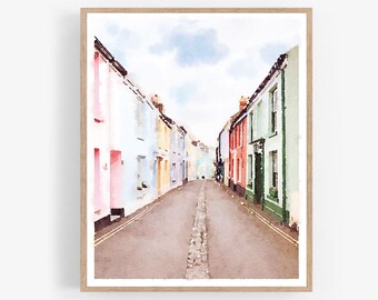 May include: Watercolor painting of a narrow street lined with colorful houses. The houses are painted in shades of pink, blue, yellow, and green. The street is paved with cobblestones and there is a light blue sky in the background.