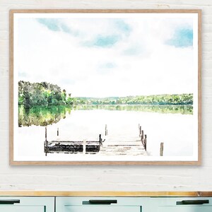 May include: Watercolor print of a serene lake scene with a wooden dock. The artwork features a light blue sky, green trees, and the reflection of the landscape in the water. The print is framed in a light brown wooden frame.