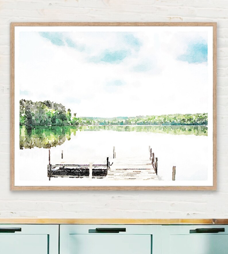 May include: Watercolor print of a serene lake scene with a wooden dock. The artwork features a light blue sky, green trees, and the reflection of the landscape in the water. The print is framed in a light brown wooden frame.