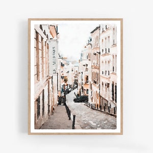 A watercolor painting of a street scene in Paris, France. The street is lined with buildings in shades of beige and white. The buildings have windows and doors, and some have signs. The street is cobblestone and there are trees and plants on either side. The sky is a light blue color. Paris Art Prints Digital Download Wall Art Set of 3, Paris Photography, Neutral Art, Paris Wall Decor, France Buildings Streets Watercolor