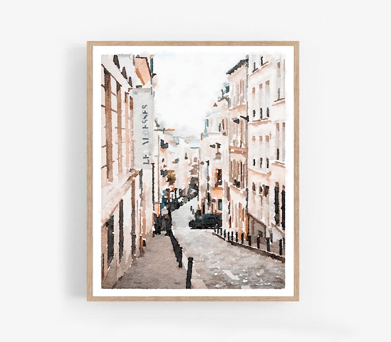 A watercolor painting of a street scene in Paris, France. The street is lined with buildings in shades of beige and white. The buildings have windows and doors, and some have signs. The street is cobblestone and there are trees and plants on either side. The sky is a light blue color. Paris Art Prints Digital Download Wall Art Set of 3, Paris Photography, Neutral Art, Paris Wall Decor, France Buildings Streets Watercolor