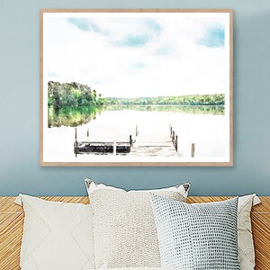 May include: Watercolor painting of a serene lake scene with a wooden dock extending into the water. The artwork features a light blue sky with clouds, green trees, and their reflection in the calm water. The painting is framed in a light brown wooden frame.