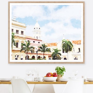 Pode incluir: Impress&atilde;o em aquarela de uma paisagem urbana com edif&iacute;cios brancos, telhados de telha vermelha e palmeiras. A obra de arte &eacute; emoldurada em uma moldura de madeira clara. O c&eacute;u &eacute; azul claro com nuvens brancas. A impress&atilde;o &eacute; exibida em uma mesa de jantar.