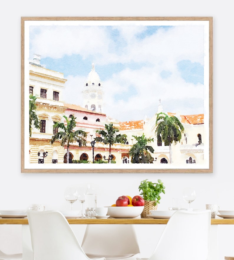 Pode incluir: Impress&atilde;o em aquarela de uma paisagem urbana com edif&iacute;cios brancos, telhados de telha vermelha e palmeiras. A obra de arte &eacute; emoldurada em uma moldura de madeira clara. O c&eacute;u &eacute; azul claro com nuvens brancas. A impress&atilde;o &eacute; exibida em uma mesa de jantar.