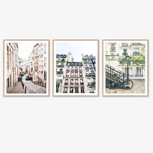 Paris Art Prints Digital Download Wall Art Set of 3, Paris Photography, Neutral Art, Paris Wall Decor, France Buildings Streets Watercolor. A set of three watercolor paintings depicting Parisian streets. The first painting shows a narrow street with buildings on both sides. The second painting shows a close-up of a building with windows and balconies. The third painting shows a bicycle parked on a cobblestone street with a lamppost and greenery in the background.