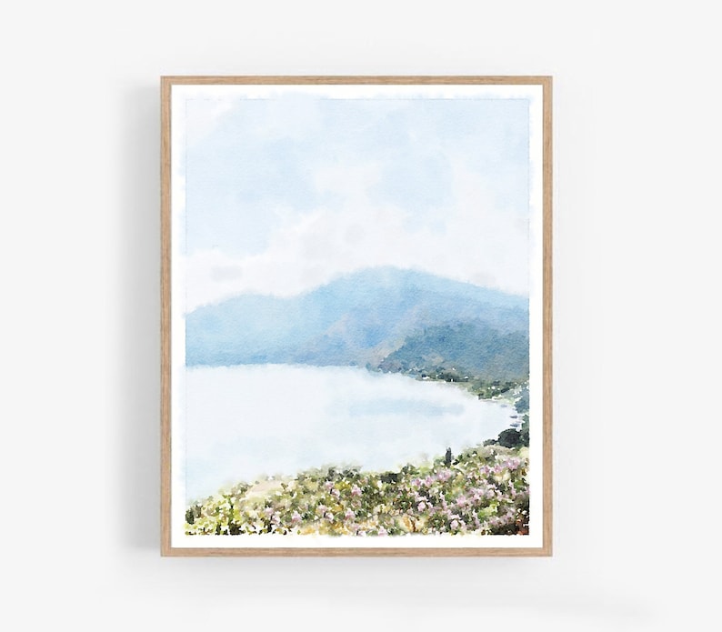 May include: A watercolor painting of a mountain range and a lake with a light blue sky and white clouds. The lake is surrounded by green trees and bushes. The painting is framed in a light wood frame.