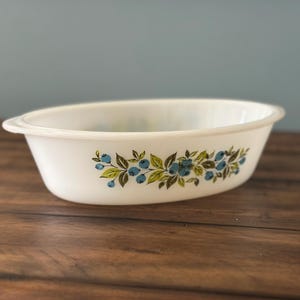 Pode incluir: Uma assadeira oval de vidro leitoso branco com um padrão decorativo de bagas azuis e folhas verdes na lateral externa. O recipiente possui alças integradas em ambas as extremidades e um acabamento liso e brilhante em toda a superfície.