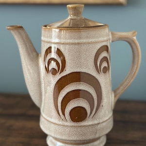 May include: A ceramic coffee pot featuring a speckled cream and brown glaze. The body is decorated with concentric brown circular patterns. It includes a matching lid with a small knob, a curved handle, and a tapered spout on the side.