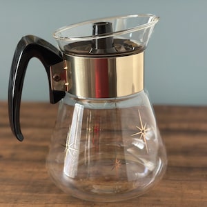 May include: A clear glass coffee carafe featuring a gold-toned metal band around the neck and a black plastic handle. The glass body is decorated with small, multi-coloured starburst patterns in red and gold. It includes a black plastic lid.