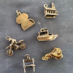 Peut inclure: Un ensemble de six pendentifs de charme en argent, comprenant un bateau, un b&acirc;timent, une moto, une chaise, une selle et un personnage de dessin anim&eacute;. Les breloques sont toutes en argent et ont une finition brillante.