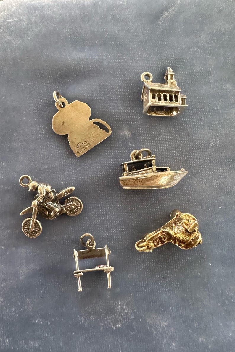 Peut inclure: Un ensemble de six pendentifs de charme en argent, comprenant un bateau, un b&acirc;timent, une moto, une chaise, une selle et un personnage de dessin anim&eacute;. Les breloques sont toutes en argent et ont une finition brillante.