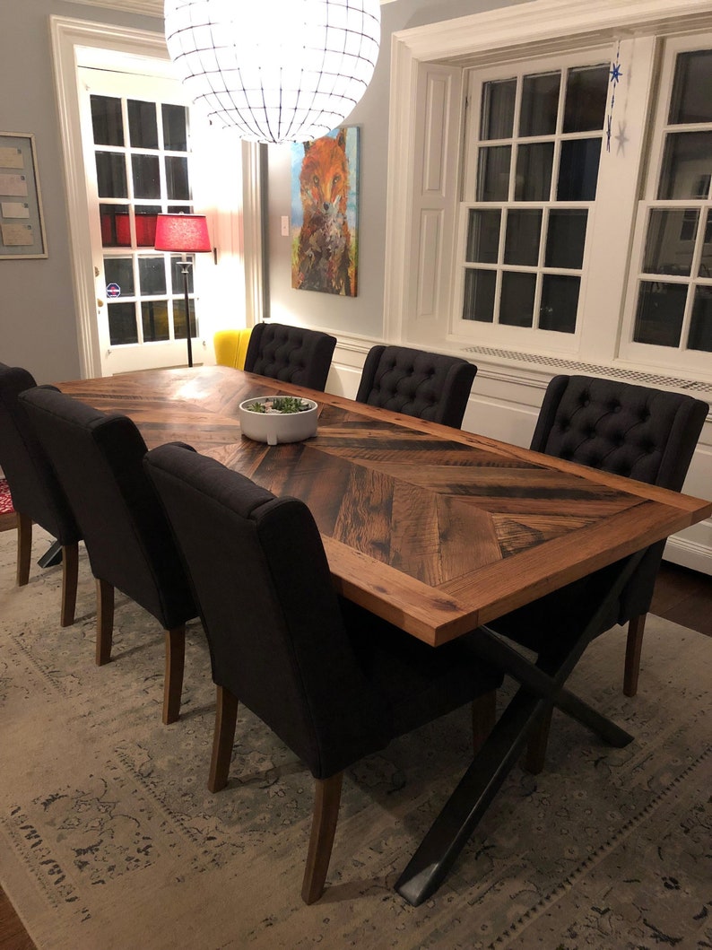Pu&ograve; includere: Un tavolo da pranzo in legno rustico con un piano a motivo chevron e gambe in metallo nero a forma di X. Il tavolo &egrave; circondato da sei sedie imbottite nere.