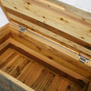 Puede incluir: Un cofre de almacenamiento de madera con tapa con bisagras, hecho de madera recuperada con una variedad de tonos y texturas de madera. El cofre est&aacute; abierto, revelando el interior, que tambi&eacute;n est&aacute; hecho de madera recuperada.
