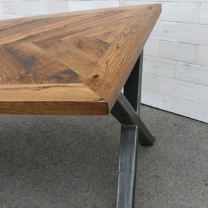 Pu&ograve; includere: Un tavolo in legno con un piano a motivo chevron e gambe in metallo nero. Il tavolo &egrave; realizzato in legno riciclato e ha un aspetto rustico.