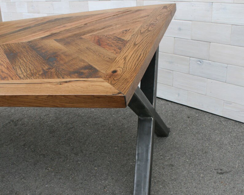Pu&ograve; includere: Un tavolo in legno con un piano a motivo chevron e gambe in metallo nero. Il tavolo &egrave; realizzato in legno riciclato e ha un aspetto rustico.