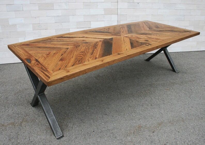 Pu&ograve; includere: Un lungo tavolo in legno con un piano a motivo chevron e gambe in metallo nero. Il tavolo &egrave; realizzato in legno riciclato e ha un aspetto rustico.