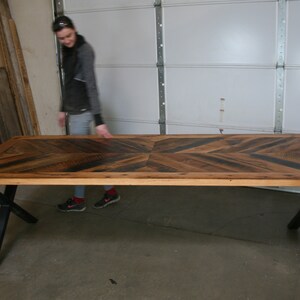 Pu&ograve; includere: Un grande tavolo in legno con un piano a chevron e gambe a X in metallo nero. Il tavolo &egrave; realizzato in legno riciclato e ha un aspetto rustico.
