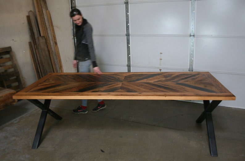 Pu&ograve; includere: Un grande tavolo in legno con un piano a chevron e gambe a X in metallo nero. Il tavolo &egrave; realizzato in legno riciclato e ha un aspetto rustico.