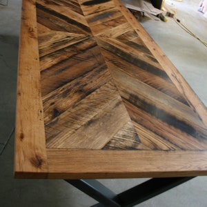 Pu&ograve; includere: Un piano da tavolo in legno rustico con un motivo a chevron realizzato in legno riciclato. Il piano del tavolo ha una finitura in legno naturale con una macchia marrone scuro. Il piano del tavolo &egrave; circondato da una cornice in legno massiccio spessa. Il piano del tavolo &egrave; sostenuto da una base in metallo nero a forma di X.