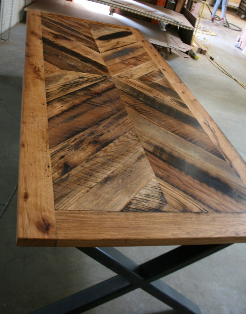 Pu&ograve; includere: Un piano da tavolo in legno rustico con un motivo a chevron realizzato in legno riciclato. Il piano del tavolo ha una finitura in legno naturale con una macchia marrone scuro. Il piano del tavolo &egrave; circondato da una cornice in legno massiccio spessa. Il piano del tavolo &egrave; sostenuto da una base in metallo nero a forma di X.