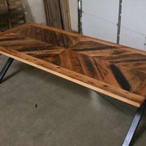 Pu&ograve; includere: Un tavolo da pranzo in legno rustico con un piano a chevron e gambe in metallo nero. Il tavolo &egrave; realizzato in legno riciclato e ha una finitura naturale.