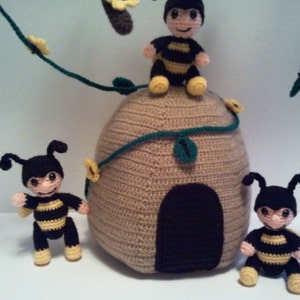 Pu&ograve; includere: Decorazioni per la cameretta a tema api all'uncinetto con un alveare beige con un ingresso marrone scuro, circondato da diverse api nere e gialle. Le api hanno facce sorridenti e sono posizionate attorno all'alveare, con un tralcio di foglie verdi e fiori gialli.