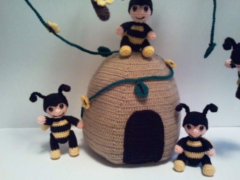 Pu&ograve; includere: Decorazioni per la cameretta a tema api all'uncinetto con un alveare beige con un ingresso marrone scuro, circondato da diverse api nere e gialle. Le api hanno facce sorridenti e sono posizionate attorno all'alveare, con un tralcio di foglie verdi e fiori gialli.