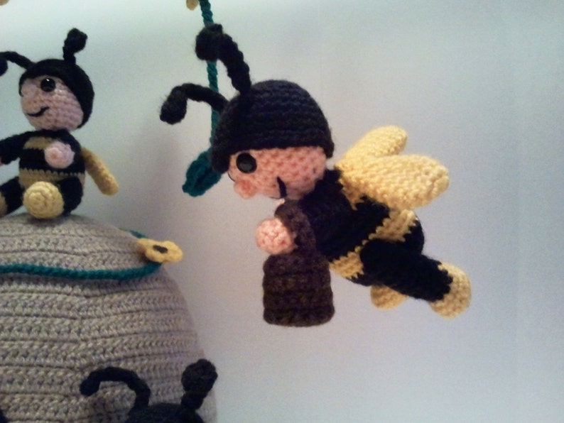 Pu&ograve; includere: Mobile a tema api realizzato all'uncinetto, con un alveare grigio con bordi turchesi e diverse api nere e gialle. Un'ape &egrave; in volo e un'altra &egrave; appollaiata sull'alveare. Le api hanno antenne e cappelli neri.