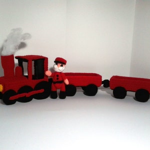Puede incluir: Un tren de juguete de crochet rojo con una bocanada de humo blanco saliendo de la chimenea. El tren tiene una figura de conductor de pie en la locomotora. El tren tiene dos vagones.