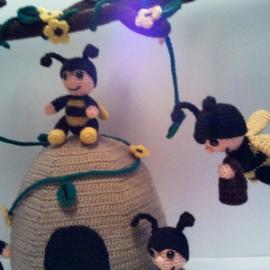 Pu&ograve; includere: Un'eccentrica giostrina all'uncinetto con diverse figure di api sorridenti. Le api hanno corpi a strisce nere e gialle, berretti neri e ali gialle. Sono posizionate attorno a un alveare beige, con un ramo marrone sopra adornato con fiori gialli e foglie verdi.