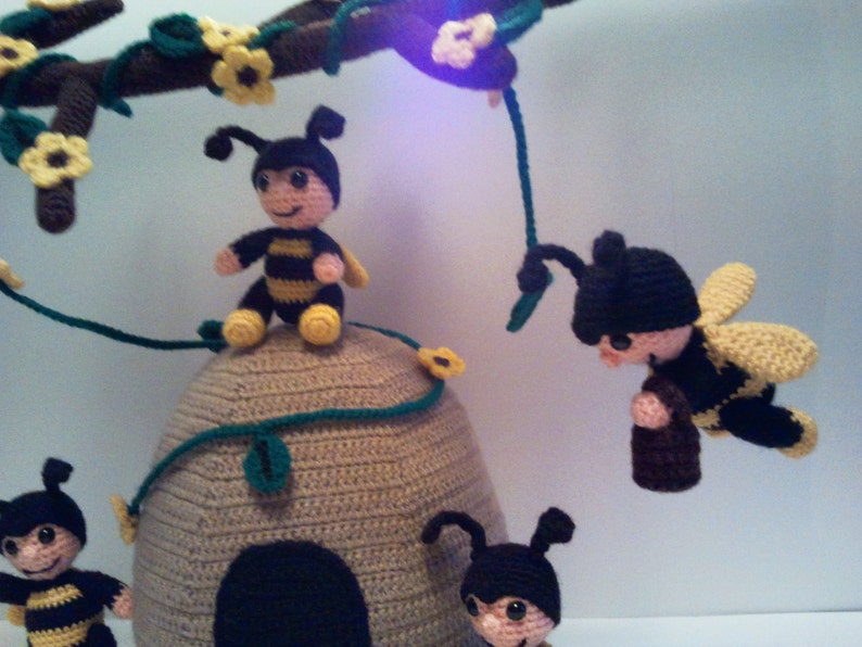 Pu&ograve; includere: Un'eccentrica giostrina all'uncinetto con diverse figure di api sorridenti. Le api hanno corpi a strisce nere e gialle, berretti neri e ali gialle. Sono posizionate attorno a un alveare beige, con un ramo marrone sopra adornato con fiori gialli e foglie verdi.