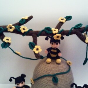Pu&ograve; includere: Mobile per la cameretta a tema api realizzato all'uncinetto. Presenta un ramo marrone decorato con fiori gialli e foglie verdi. Sono incluse diverse piccole figure di api e un alveare, tutti in filato.