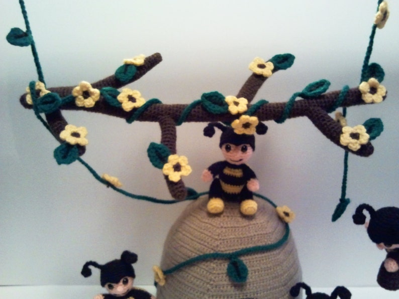 Pu&ograve; includere: Mobile per la cameretta a tema api realizzato all'uncinetto. Presenta un ramo marrone decorato con fiori gialli e foglie verdi. Sono incluse diverse piccole figure di api e un alveare, tutti in filato.