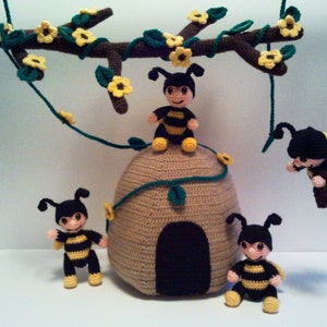 Pu&ograve; includere: Scena all'uncinetto con un alveare e diverse piccole figure di api. Le api sono nere e gialle, con facce sorridenti. Un ramo all'uncinetto con fiori gialli e foglie verdi &egrave; appeso sopra l'alveare.