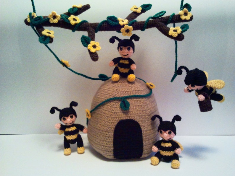 Pu&ograve; includere: Scena all'uncinetto con un alveare e diverse piccole figure di api. Le api sono nere e gialle, con facce sorridenti. Un ramo all'uncinetto con fiori gialli e foglie verdi &egrave; appeso sopra l'alveare.