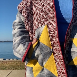 Puede incluir: Chaqueta acolchada con un patr&oacute;n geom&eacute;trico en burdeos y blanco, con detalles grises y amarillos en forma de estrella. La chaqueta tiene un ribete rojo y se usa sobre una camiseta azul y vaqueros.