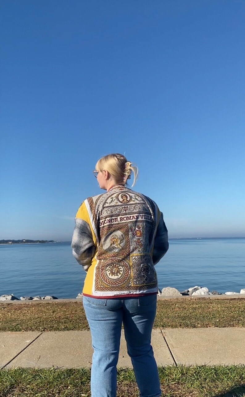 Puede incluir: Una chaqueta con un dise&ntilde;o decorativo y el texto "BIGNOR ROMAN VILLA". La chaqueta tiene un estampado de cuadros amarillos y grises en las mangas y la espalda. La persona lleva vaqueros azules.