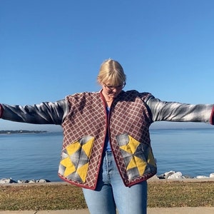 Puede incluir: Chaqueta acolchada con un estampado geom&eacute;trico en rojo, gris y amarillo. La chaqueta tiene un ribete burdeos y se lleva con vaqueros azules. El fondo presenta una masa de agua y un cielo azul claro.