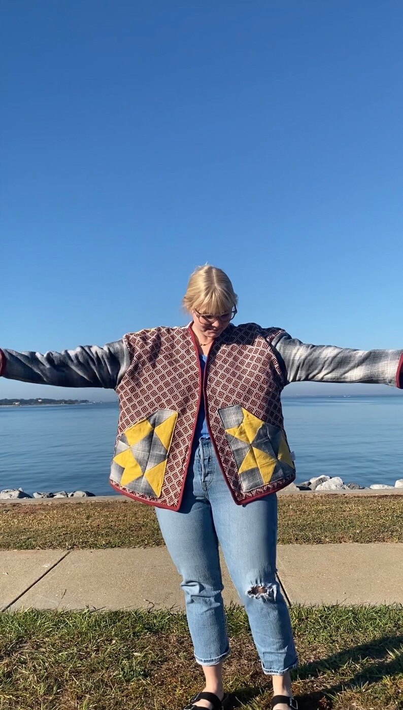 Puede incluir: Chaqueta acolchada con un estampado geom&eacute;trico en rojo, gris y amarillo. La chaqueta tiene un ribete burdeos y se lleva con vaqueros azules. El fondo presenta una masa de agua y un cielo azul claro.