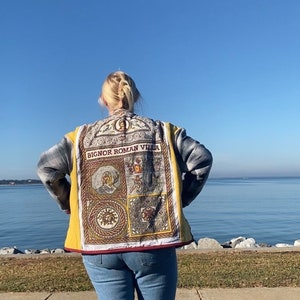 Puede incluir: Chaqueta acolchada amarilla y gris con un panel decorativo con el texto "BIGNOR ROMAN VILLA" y varias im&aacute;genes de tem&aacute;tica romana. La chaqueta se lleva sobre unos vaqueros azules. El fondo muestra una masa de agua y un cielo azul claro.