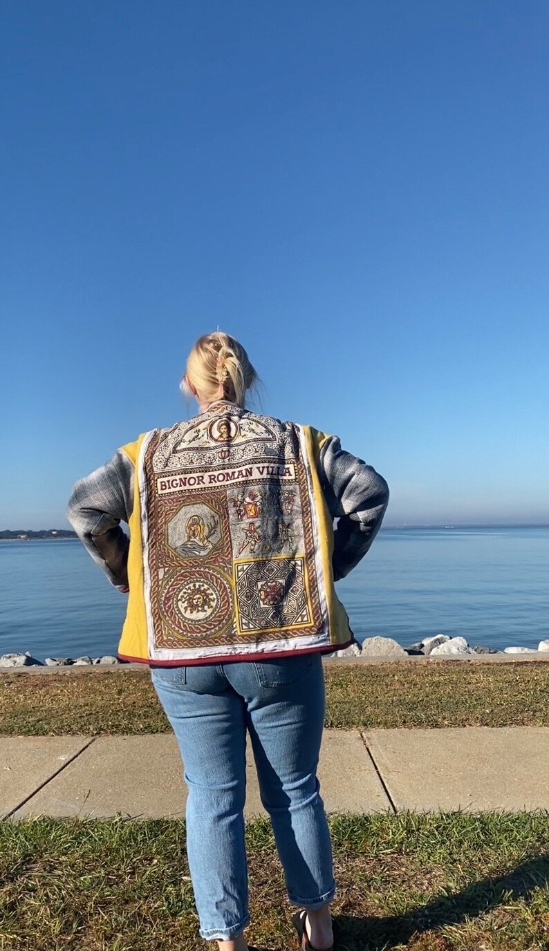 Puede incluir: Chaqueta acolchada amarilla y gris con un panel decorativo con el texto "BIGNOR ROMAN VILLA" y varias im&aacute;genes de tem&aacute;tica romana. La chaqueta se lleva sobre unos vaqueros azules. El fondo muestra una masa de agua y un cielo azul claro.