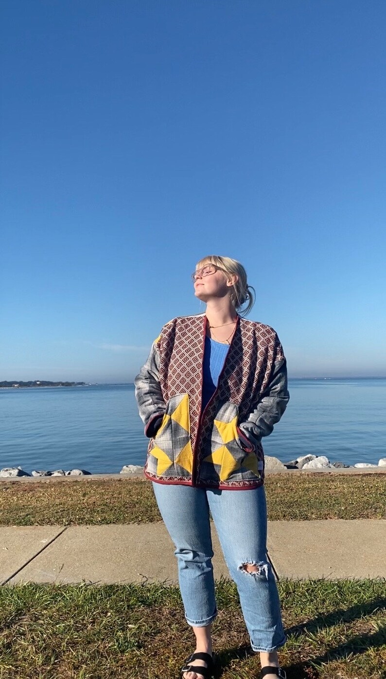 Puede incluir: Chaqueta estampada con bolsillos amarillos en forma de estrella, usada sobre una camiseta azul y vaqueros claros. La chaqueta presenta un dise&ntilde;o geom&eacute;trico en rojo, gris y marr&oacute;n. La persona lleva sandalias negras y gafas, al aire libre en un d&iacute;a soleado.