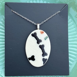 May include: Oval pendant necklace with a white background and black bone designs. The pendant is suspended from a silver chain. The pendant also has small orange and green accents. The necklace is displayed on a black card.